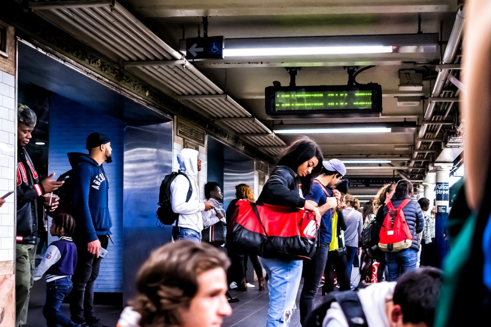 People waiting in underground transit empty large platform in NYC Subway Station in downtown, looking at phones on Fulton street