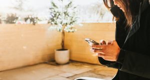 Woman sits outside in a sunny area, surrounded by a wall and a tree. She grips a mobile phone with both hands and scrolls and types with both thumbs. She seems serious / distracted / concerned as she looks down at the screen.