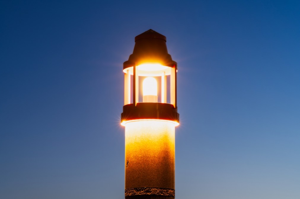The top of an illuminated lighthouse on a blue sky background at dusk.