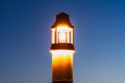 The top of an illuminated lighthouse on a blue sky background at dusk.